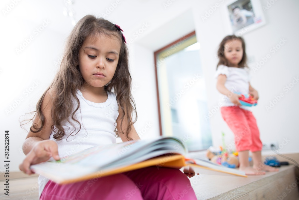 Cute girl reading book with sister playing in background at home