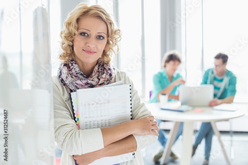 Portrait of creative businesswoman holding files with colleagues working in background at office