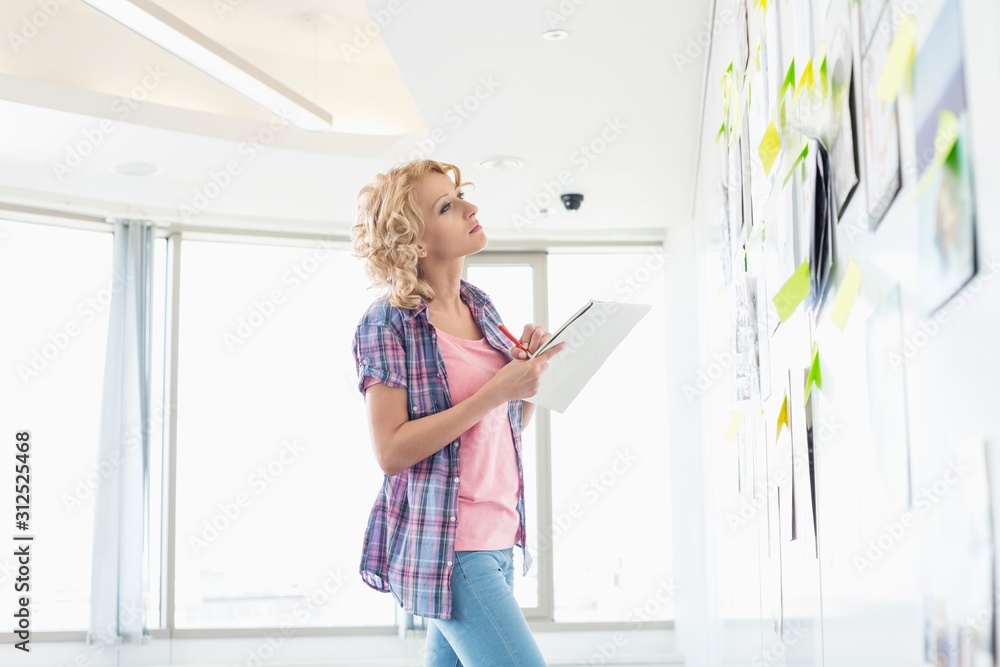 Creative businesswoman looking at papers stuck on wall while writing notes in office Creative businesswoman looking at papers stuck on wall while writing notes in office