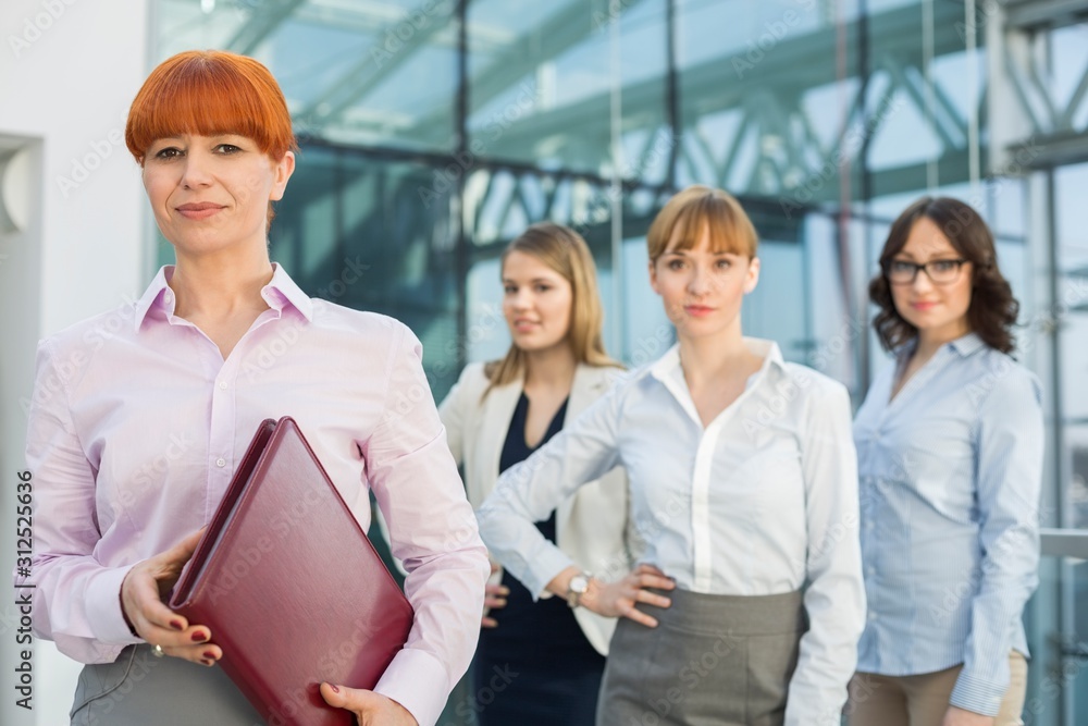 Portrait of confident businesswoman holding folder with female colleagues standing in background Portrait of confident businesswoman holding folder with female colleagues standing in background