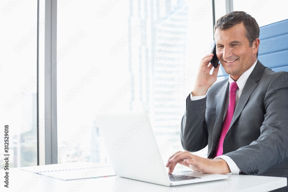 Smiling mature businessman talking on cell phone while using laptop in office Smiling mature businessman talking on cell phone while using laptop in office