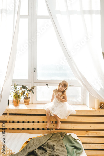 Cute little girl in beautiful dress is sitting on a window sill at light sunny bedroom