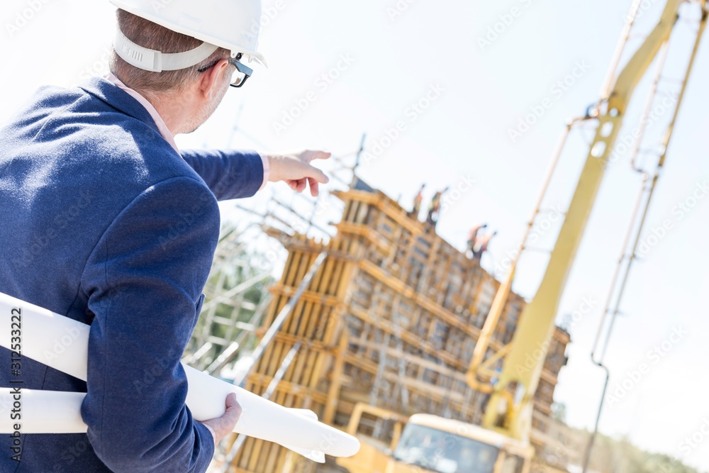 Rear view of architect holding blueprints while pointing at construction site Rear view of architect holding blueprints while pointing at construction site