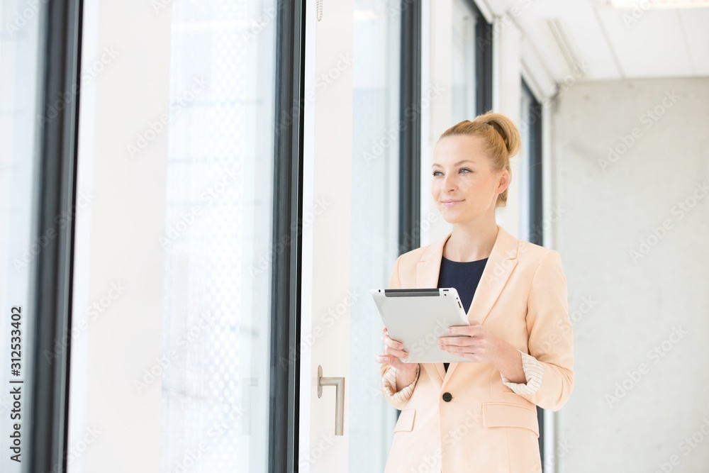 Young businesswoman holding digital tablet while looking away in office Young businesswoman holding digital tablet while looking away in office