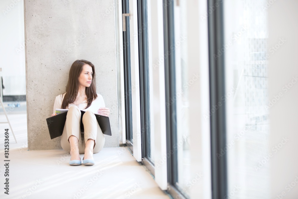 Young businesswoman with file looking through office window while sitting on floor