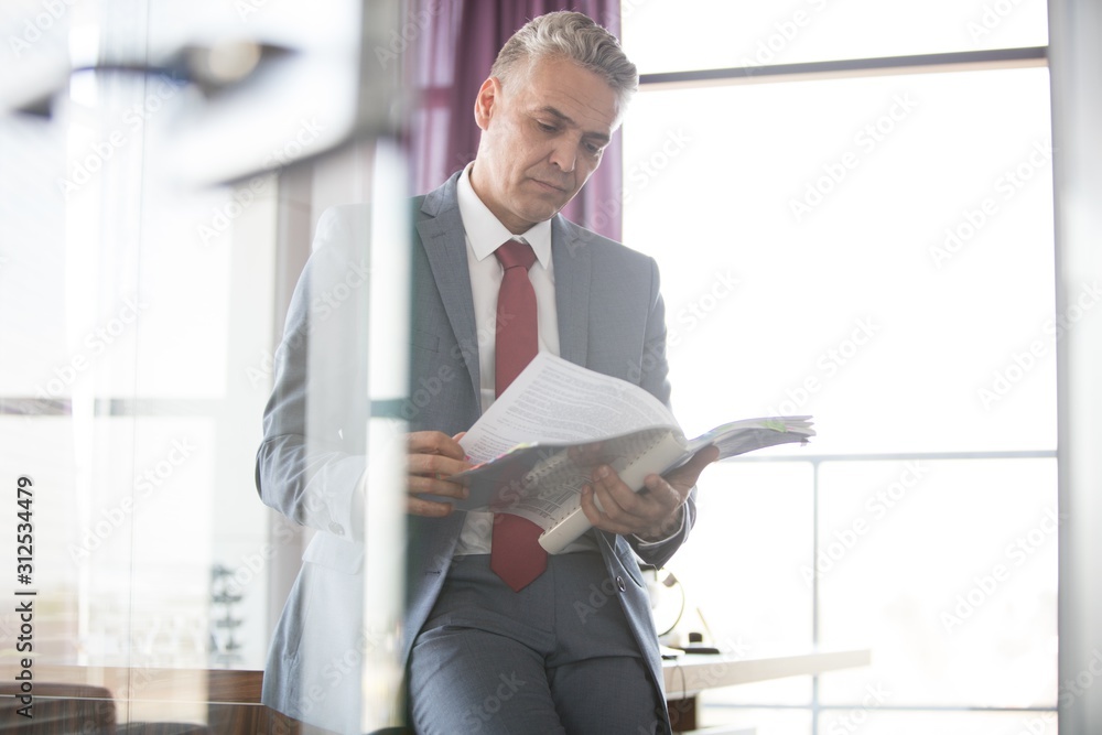 Fototapeta premium Mature businessman reading book while leaning on desk in office