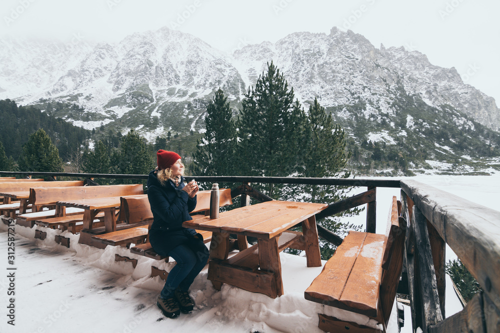 Woman having a hot drink from thermo cup overlooking snowcapped High Tatra mountains peaks