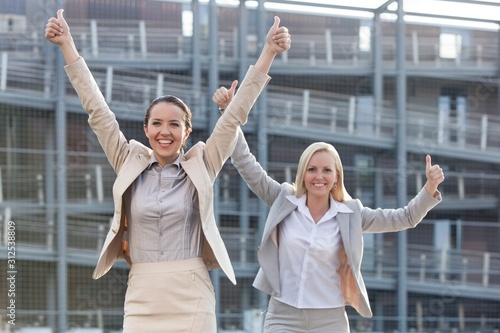 Excited young businesswomen gesturing thumbs up against office building