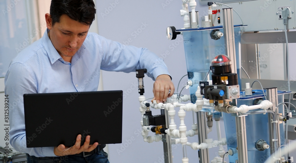 Focus on Engineer who is programming PLC with laptop for an analog process kit. Industry 4.0 workshop in an automation education center. Engineer is learning how to program a PLC. Focus on Engineer who is programming PLC with laptop for an analog process kit. Industry 4.0 workshop in an automation education center. Engineer is learning how to program a PLC.