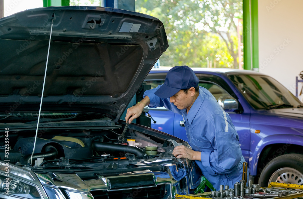 Asian automotive mechanic with blue uniform use wrench to fix the problem in part of engine around bonnet and front bumper of car in garage. Asian automotive mechanic with blue uniform use wrench to fix the problem in part of engine around bonnet and front bumper of car in garage.