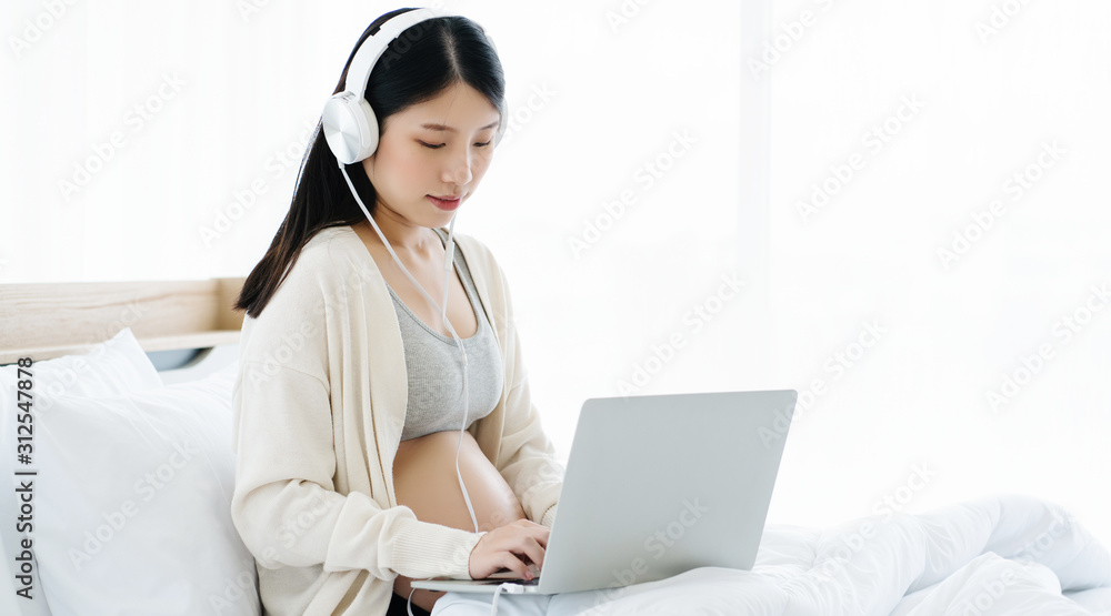 Beautiful Asian pregnant women wear headphones, use a laptop, find information about the health care of the mother and the child in the womb. Sitting on clean bed in a bedroom with a good environment Beautiful Asian pregnant women wear headphones, use a laptop, find information about the health care of the mother and the child in the womb. Sitting on clean bed in a bedroom with a good environment