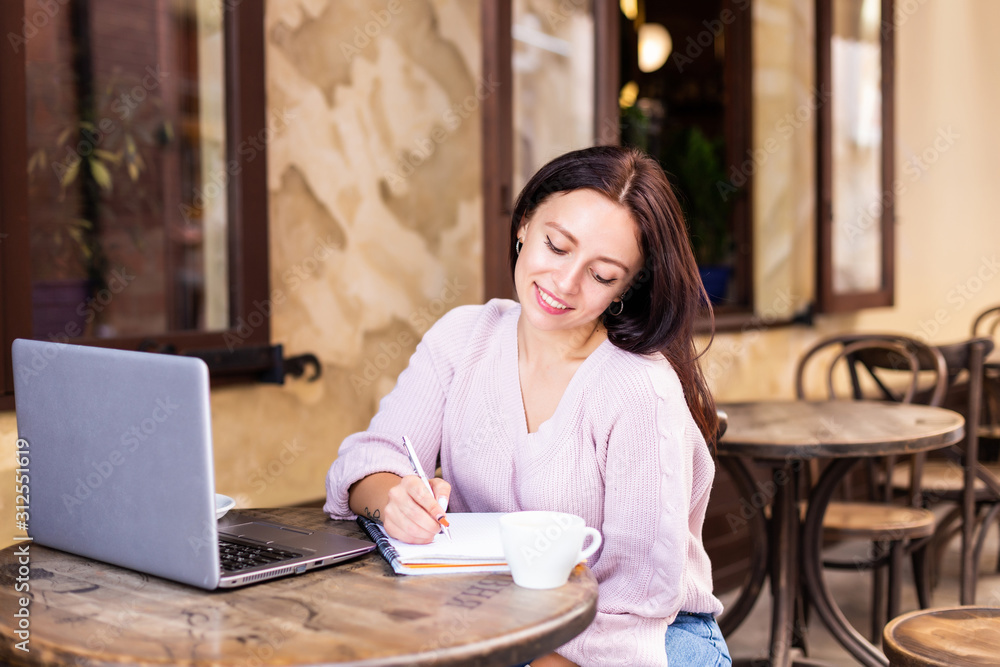 Smiling woman in cafe using laptop and note some data on notebook Smiling woman in cafe using laptop and note some data on notebook