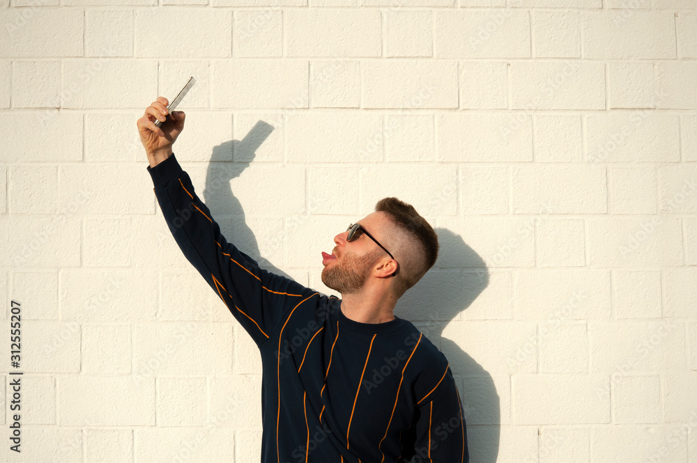 Positive smiling guy in casualwear showing tongue with mobile phone taking selfie. Young bearded man standing at outdoor white wall. Male selfie, leisure, fun concept. Positive smiling guy in casualwear showing tongue with mobile phone taking selfie. Young bearded man standing at outdoor white wall. Male selfie, leisure, fun concept.