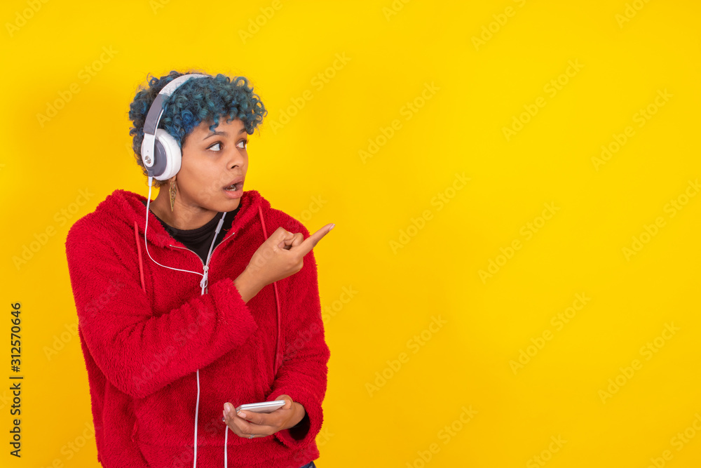 young african American woman with mobile phone and headphones pointing astonished isolated on yellow background young african American woman with mobile phone and headphones pointing astonished isolated on yellow background