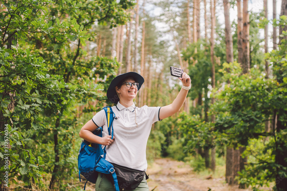 Smiling female tourist holds smart phone, takes selfie against beautiful landscape wood. Hiking woman with backpack taking selfie photo with smartphone. Travel and healthy lifestyle outdoors