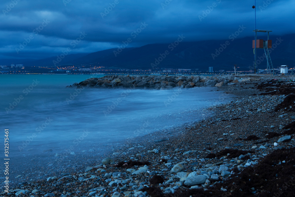 Sea surf on a long exposure in the evening. Waves roll over the pebbly beach and rocks. In the background, mountains and city lights. 
