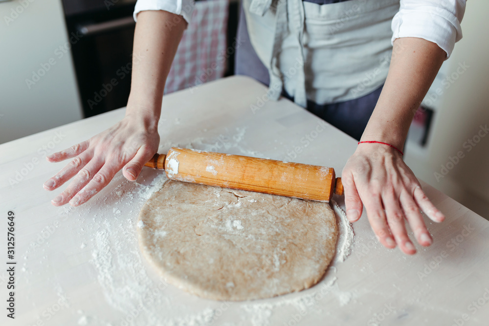 Crop woman rolls out the dough with a wooden rolling pin on the table for cooking.