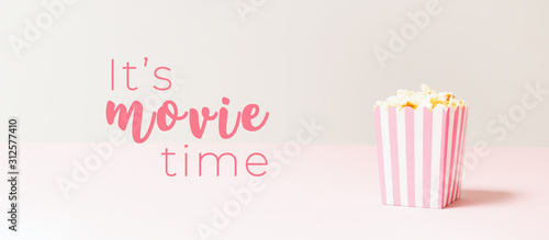 Popcorn bag with white and pink stripes at the light neutral backdrop. Empty minimalistic background. Romantic movie night and snack concept with It s movie time wording. Banner wide screen format