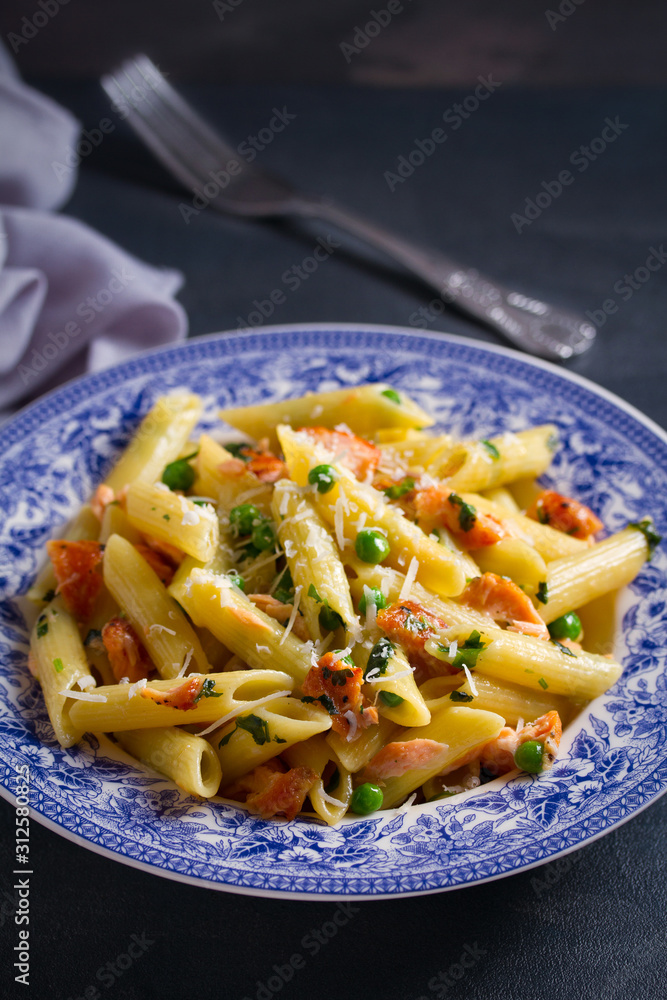 Creamy salmon penne pasta with parmesan cheese green peas in blue bowl on gray background