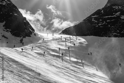 Silhouettes of many skiers in the back light in the afternoon between two mountain peaks on the ski piste from Porta Vescovo to Arabba, Italy. Stormy weather and snow dust in the air; black and white photo