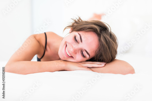 A woman lying at the end of the bed underneath the quilt and smiling  with her head resting upon her hand.