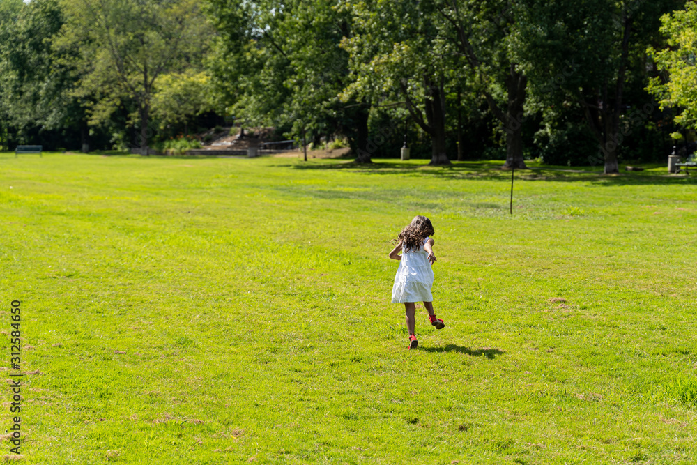 back view of brunnette young girl with white dress and red shoes running in green field near a forest, sun shining in spring