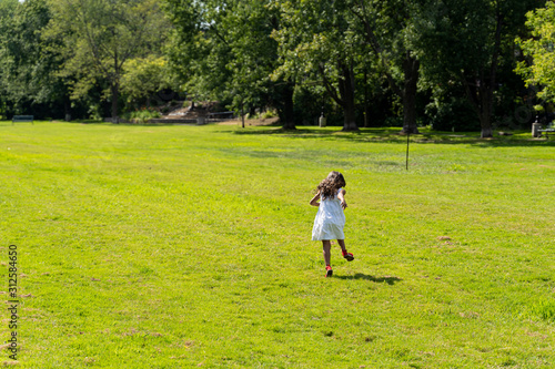 back view of brunnette young girl with white dress and red shoes running in green field near a forest  sun shining in spring
