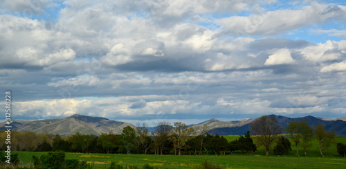 wide shot of blue skies and white fluffy clouds flowing over a mountain side with green pastures in the foreground
