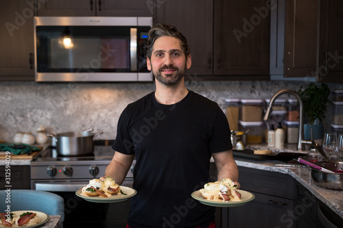 A ethnically mixed male homecook in a kitchen holding plates of eggs benedict. photo