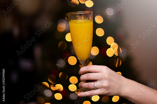 A womans hand holding a mimosa in front of a christmas tree to celebrate the holidays.