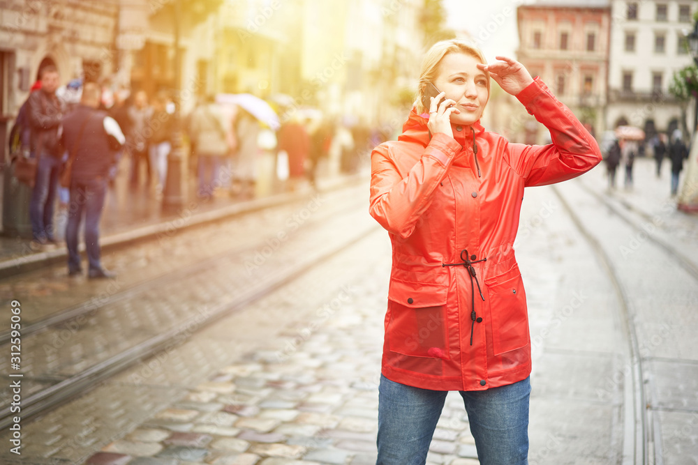 Girl talking on the phone on the street. Portrait of pretty smiling young woman in red bright raincoat. Sunny raining day in city. Image with flare .