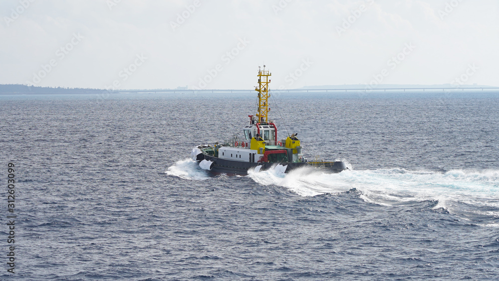 Fototapeta premium Colored tugboat works in port. bright tugboat in blue water. industrial ship on waves tropical islands. powerful tugboat helps maneuvering big ship turquoise blue Pacific Ocean Okinawa Japan