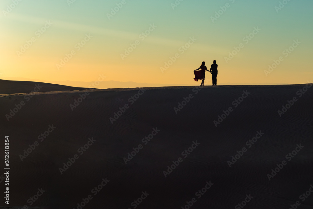 A couple on a desert dune in silhouette at sunset with the woman holding her dress.