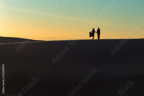 A couple on a desert dune in silhouette at sunset with the woman holding her dress. © sherryvsmith