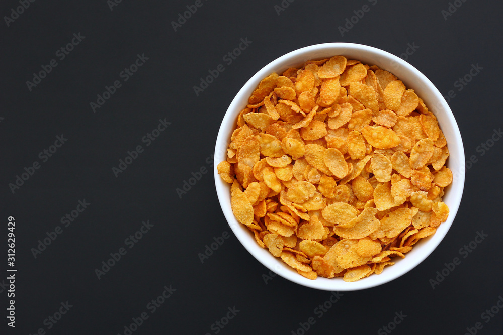 Dry corn flakes in a white bowl on a black metallic surface. Copy space. Dry corn flakes in a white bowl on a black metallic surface. Copy space.