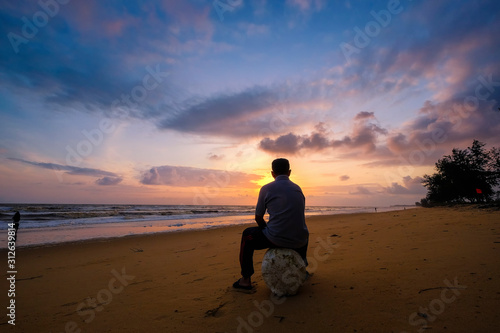 Unidentified man sitting with soft wave of blue ocean on sandy beach during sunrise