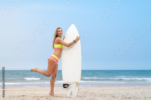 Blonde surfer woman in a bikini with a white surfboard standing on the sand beach looking at the blue sea on a surf spot in Vietnam Asia