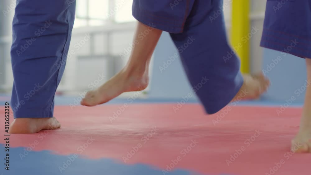 Low section shot in slow motion of barefoot female jujutsu athlete doing cling leg trip takedown and throwing male partner down on mat during training session Low section shot in slow motion of barefoot female jujutsu athlete doing cling leg trip takedown and throwing male partner down on mat during training session