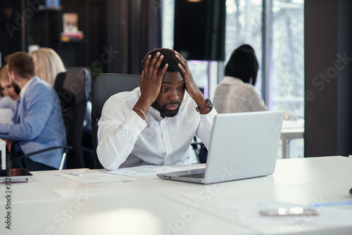 Disappointed african american office manager looking on the laptop screen and put his hands on head at his work table on the coworkers background.