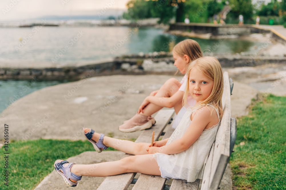 Little girls playing by the lake in summertime, children having fun outside, resting on the bench