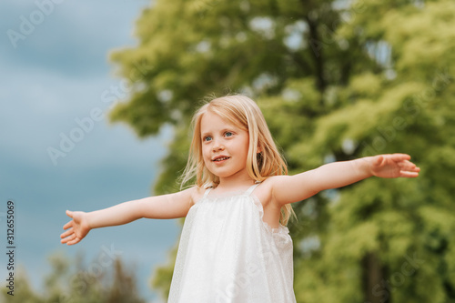 Outdoor portrait of cute little girl with blond hair, arms wide open