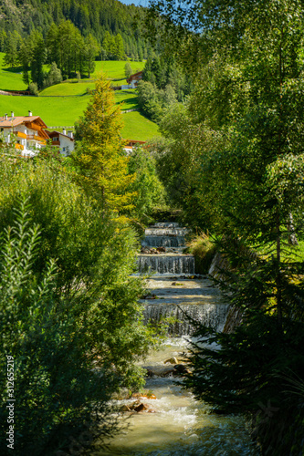 mountain stream descends from the dolomites with a small waterfall and stone wall among lush green trees