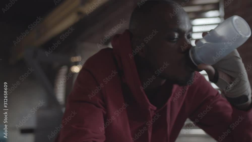 Chest-up shot of tired young black fighter in hooded sweatshirt, with sweaty face leaning on ropes in boxing ring at fight club, drinking water from bottle and resting Chest-up shot of tired young black fighter in hooded sweatshirt, with sweaty face leaning on ropes in boxing ring at fight club, drinking water from bottle and resting