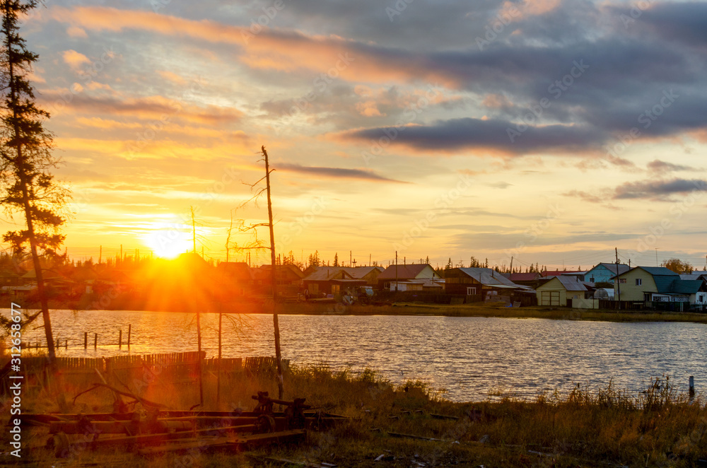 Bright colorful sunset in the village of Yakutia Suntar on the background of old fir trees by the lake with houses on the shore under a cloudy sky.