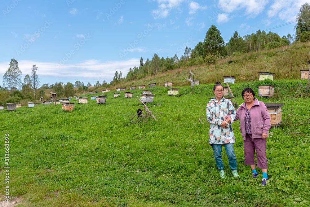 An Asian Yakut family an elderly mother and a young daughter pose hand in hand against the background of hives for bees in an apiary in the Altai mountains. An Asian Yakut family an elderly mother and a young daughter pose hand in hand against the background of hives for bees in an apiary in the Altai mountains.