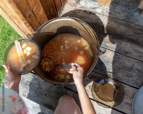 Girl in nature puts in a bowl of soup borscht with meat and bone and vegetables from a large pot on a wooden floor bright day for lunch in the wild.