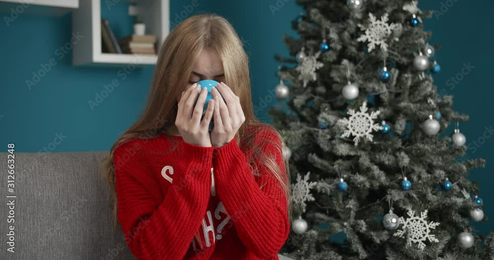 Cute smiling lady in red sweater drinking coffee and looking to camera near decorated Christmas tree