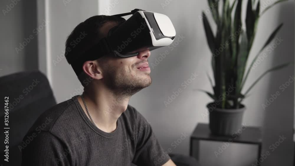 Young bearded man wearing headset of virtual reality in office sitting on a sofa. Exploring VR world and all what he saw in headset. Young bearded man wearing headset of virtual reality in office sitting on a sofa. Exploring VR world and all what he saw in headset.
