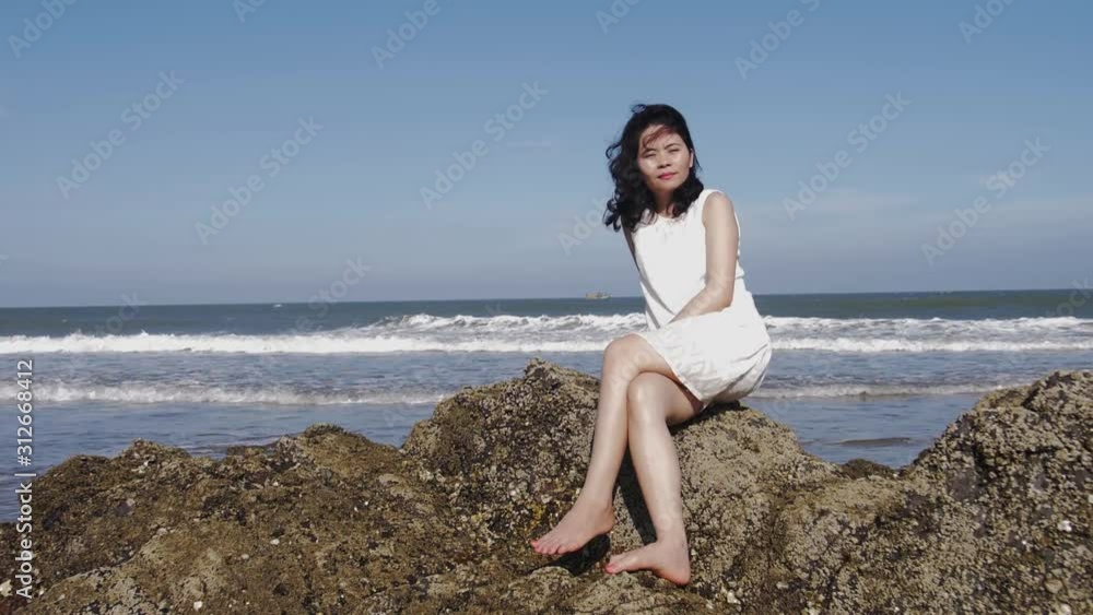 Dark-haired woman looks toward camera while sitting on rock by ocean in Vietnam, slomo Dark-haired woman looks toward camera while sitting on rock by ocean in Vietnam, slomo
