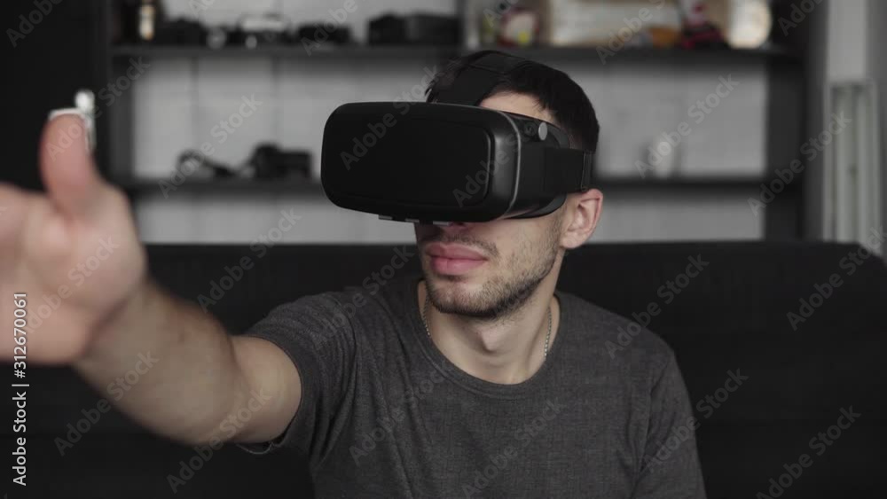 Young bearded man wearing headset of virtual reality in office sitting on a sofa and trying to touch something that he see in virtual world. Young bearded man wearing headset of virtual reality in office sitting on a sofa and trying to touch something that he see in virtual world.